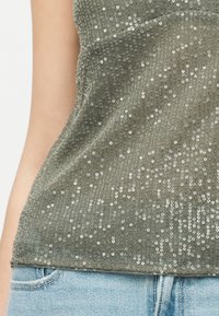 Κοντινό πλάνο ενός πράσινου τοπάκι με παγιέτες που φοριέται με ανοιχτόχρωμο μπλε τζιν, δείχνοντας τις λεπτομέρειες υφής και λάμψης στο ύφασμα.