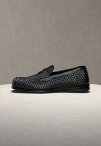 Mocassin en cuir noir avec motif tissé et finition élégante, présenté sur une surface claire sur fond uni.