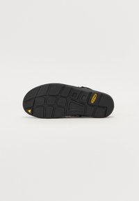 Semelle de chaussure noire en caoutchouc avec un motif de grip texturé, dotée d'un accent triangulaire jaune et d'un détail en relief "CONTOUR ARCH".