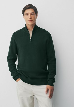 Suéter de color verde oscuro con un cuello alto y cremallera de cuarto. Puños y dobladillo acanalados; textura suave. Lleva puesto sobre una camisa clara, con las manos en los bolsillos.