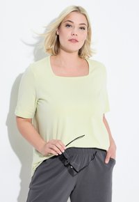 Top corto giallo chiaro con scollatura quadrata, abbinato a pantaloni grigi scuri. La modella tiene in mano occhiali da sole neri, i capelli cadono morbidamente sulle spalle.