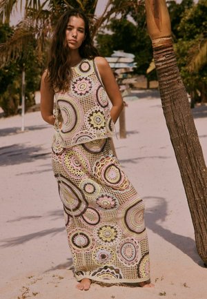 Tânără stând desculță pe nisip, purtând un top fără mâneci din croșetat și o fustă lungă cu modele circulare în tonuri pastelate, lângă palmieri.