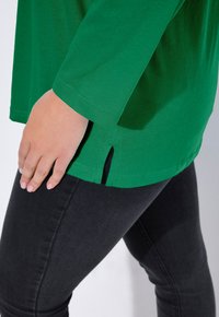 Zielona koszula z długim rękawem z zaokrąglonym dołem i bocznymi rozcięciami, wykonana z miękkiej tkaniny, zestawiona z ciemnymi jeansami o wąskim kroju. Zbliżenie na dłoń na koszuli.