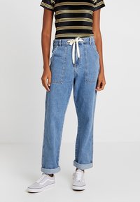 Jeans w wysokim stanem w niebieskim kolorze, luźny krój, widoczne przednie kieszenie oraz białe sznurki. Rolowane mankiety i gładka struktura.