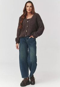 Brązowy puszysty kardigan z dużymi guzikami, zestawiony z niebieskimi spodniami typu wide-leg. Model nosi ciemne buty, prezentując swobodny i luźny styl.