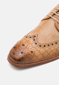 Scarpe da uomo in pelle color tan con punta affusolata, design perforato, dettagli in motivo coccodrillo e borchie nere. Texture liscia.