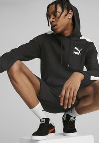 Felpa nera con cappuccio e dettagli bianchi, abbinata a short neri. Il modello indossa sneakers nere con dettagli arancioni e calze bianche.