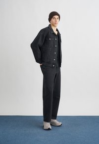 Casaco de ganga preto e calças de ganga com corte largo. Camisola preta de gola redonda por baixo. Gorro de malha castanho e ténis desportivos cinza claro.