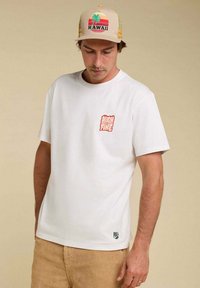 T-shirt blanc à manches courtes avec un graphique rouge "HIGH ON FIRE" sur la poitrine gauche. Matière en coton doux, avec un petit logo vert à l'ourlet.