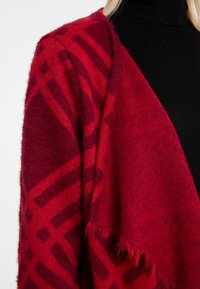 Rot und burgunderfarben gemusterter Cardigan mit einer weichen, strukturierten Oberfläche, der ein drapiertes Design und einen Fransen-Saum aufweist. Schwarzer Rollkragenpullover darunter.