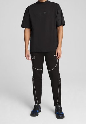 Mand iført sort oversized t-shirt og sorte Puma Motorsport bukser med hvide piping og sorte sneakers med blå accenter på grå baggrund.