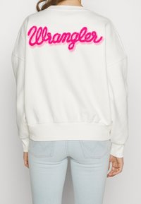 Vit sweatshirt med rosa "Wrangler"-bokstäver på ryggen, som har en avslappnad passform, ribbade muddar och rund halsringning.