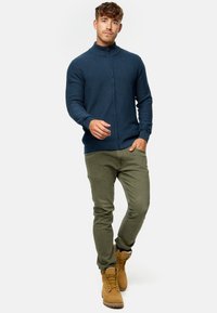 Suéter de cremallera azul marino con un cuello alto, textura de punto, combinado con pantalones verde oliva y botas marrones, que tienen un diseño con cordones y suelas resistentes.