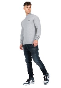Maglia grigia a maniche lunghe con collo alto, abbinata a jeans slim-fit blu scuro e sneakers alte bianco e nere. Branding evidente sulla maglietta.