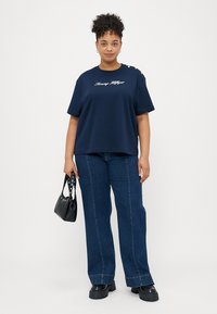 Navy Baumwoll-T-Shirt mit kurzen Ärmeln, weißem gesticktem Logo und dekorativen Schulterknöpfen. Kombiniert mit lockeren Jeans aus Denim und einer schwarzen Handtasche.