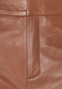 Brunt läder med en slät yta. Har en sydd söm och en subtil fickdetalj som framhäver dess skräddarsydda design.