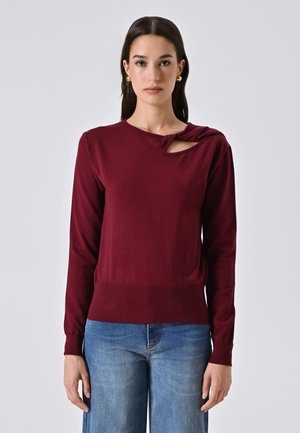 Giovane donna che indossa un maglione bordeaux a maniche lunghe con un dettaglio traforato vicino al collo, e jeans blu, in piedi di fronte a uno sfondo neutro.