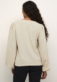 Beige strukturierte Bluse mit weitem Ausschnitt und voluminösen Ärmeln. Der Stoff hat ein dezentes, wellenförmiges Muster und wird mit dunklen Jeans kombiniert.