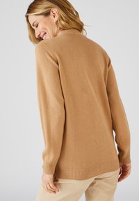 Suéter de punto marrón claro con un corte relajado y puños acanalados, que presenta un cuello redondo y una textura suave. Lleva puesto sobre unos pantalones de pana crema.