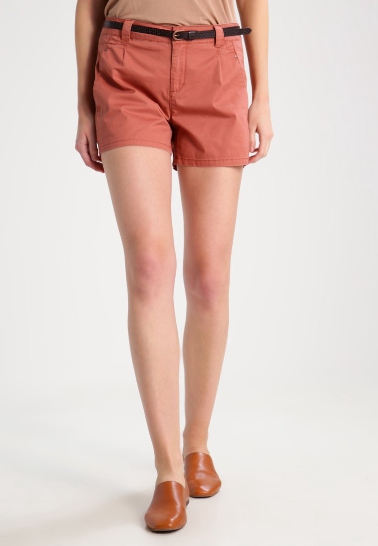 Woman wearing rust-colored shorts with black belt and brown slip-on shoes, standing against a plain light background.