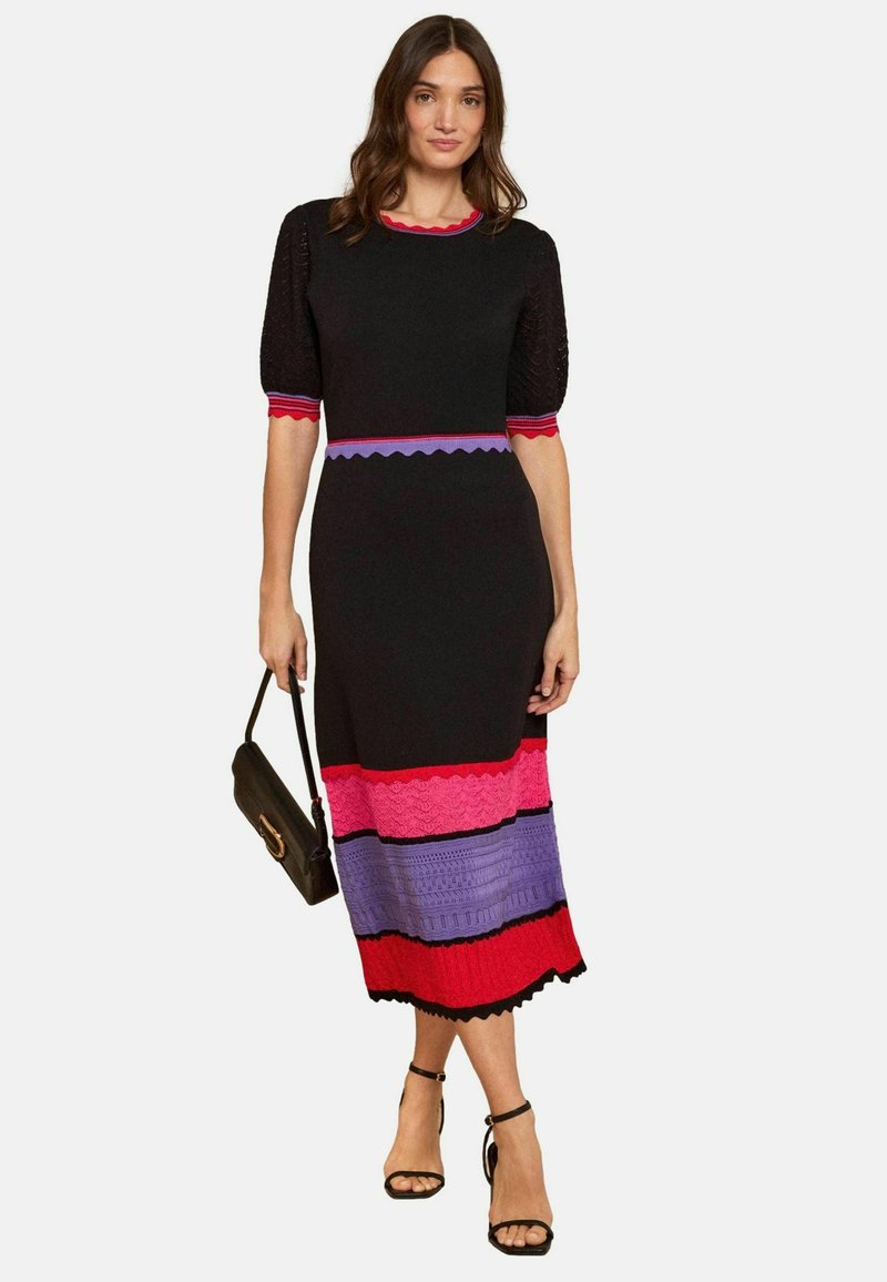 Nainen seisoo, yllään musta midi-mittainen mekko, jossa on punaisia, vaaleanpunaisia ja purppuranvärisiä vaakaraitoja, kädessä musta käsilaukku ja jalassa mustat korkokengät.