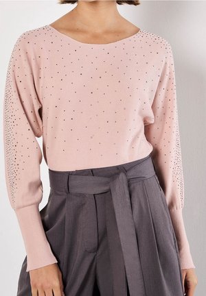 Femme portant un pull rose clair avec de petites décorations et un pantalon gris taille haute noué avec une ceinture en tissu.