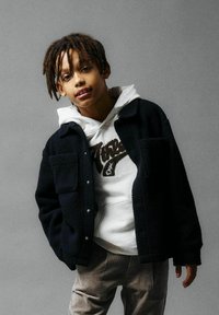 Giacca in pile nera con bottoni a pressione e due tasche anteriori, indossata sopra un cappuccio bianco con logo marrone, abbinata a pantaloni in corduroy beige.