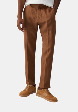 Uomo con pantaloni marroni su misura con risvolti e sneakers in camoscio color tabacco con lacci, in piedi davanti a uno sfondo semplice.