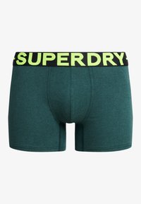 Bóxer de algodón verde con una cinturilla negra que presenta "SUPERDRY" en letras neón amarillas en negrita. Textura suave y diseño ajustado.