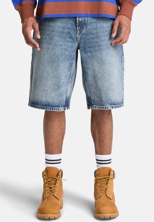 Mand iført lyseblå cowboyshorts, hvide sokker med sorte striber, brune snørebåndstøvler og en blå og brun langærmet skjorte.