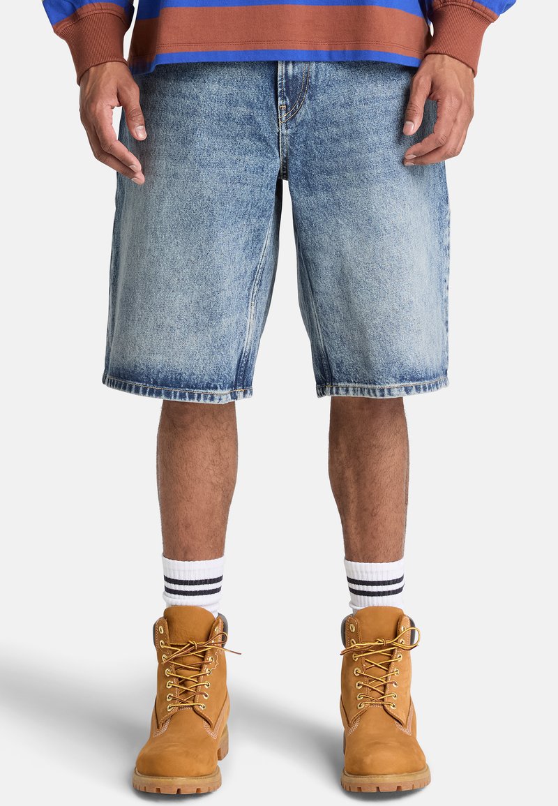 Mand iført lyseblå cowboyshorts, hvide sokker med sorte striber, brune snørebåndstøvler og en blå og brun langærmet skjorte.