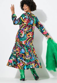 Bunt gemustertes Kleid mit gestuftem Design, das Wirbel und Augen in Neonfarben zeigt, kombiniert mit knielangen grünen Stiefeln und einem grünen Fellaccessoire.