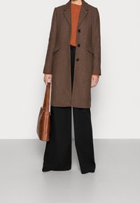 Person mit braunem Mantel, orangefarbenem Pullover, schwarzen weiten Hosen, der eine braune Lederhandtasche hält, steht auf einem hellgrauen Boden vor einer weißen Wand.