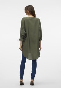 Camicia verde oliva con vestibilità rilassata, maniche lunghe e polsini arrotolati. L'orlo posteriore è leggermente più lungo, abbinata a jeans blu e tacchi.