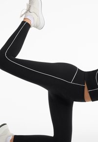 Black leggings with white side stripes, high waist design, stretchy material, fitted shape, and ankle-length cut paired with white athletic shoes.