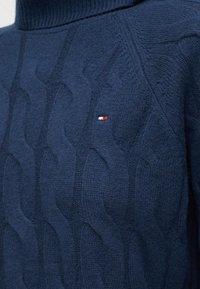 Pulover cu guler înalt, tricotat cu model în cablu, de culoare bleu marin, cu un mic logo dreptunghiular în roșu, alb și albastru pe piept.