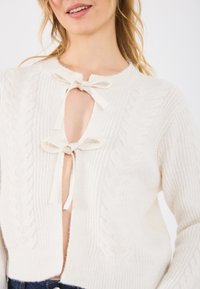 Hvit strikket cardigan med kabelmønster, en nøkkelhullfront og sløyfebåndlukking. Materialet ser mykt og teksturert ut.