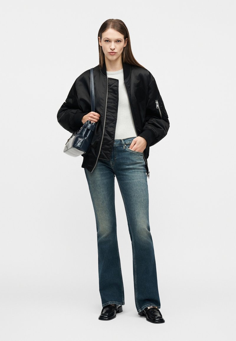 Frau trägt eine schwarze Bomberjacke, weißen Pullover, blaue ausgestellte Jeans, schwarze Loafers und eine marineblaue Handtasche über der Schulter.