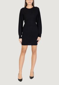 Abito nero lavorato a maglia con maniche lunghe, scollo rotondo e silhouette aderente, che arriva a metà coscia. Indossato con scarpe nere con tacco alto.