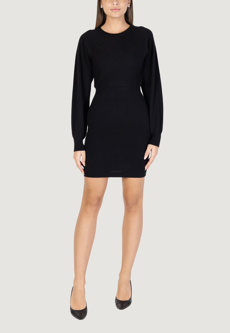 Abito nero lavorato a maglia con maniche lunghe, scollo rotondo e silhouette aderente, che arriva a metà coscia. Indossato con scarpe nere con tacco alto.