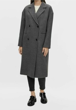 Persoon draagt een lange, donkergrijze jas met dubbele rij knopen, zwarte broek en glanzende zwarte loafers op een effen lichte achtergrond.