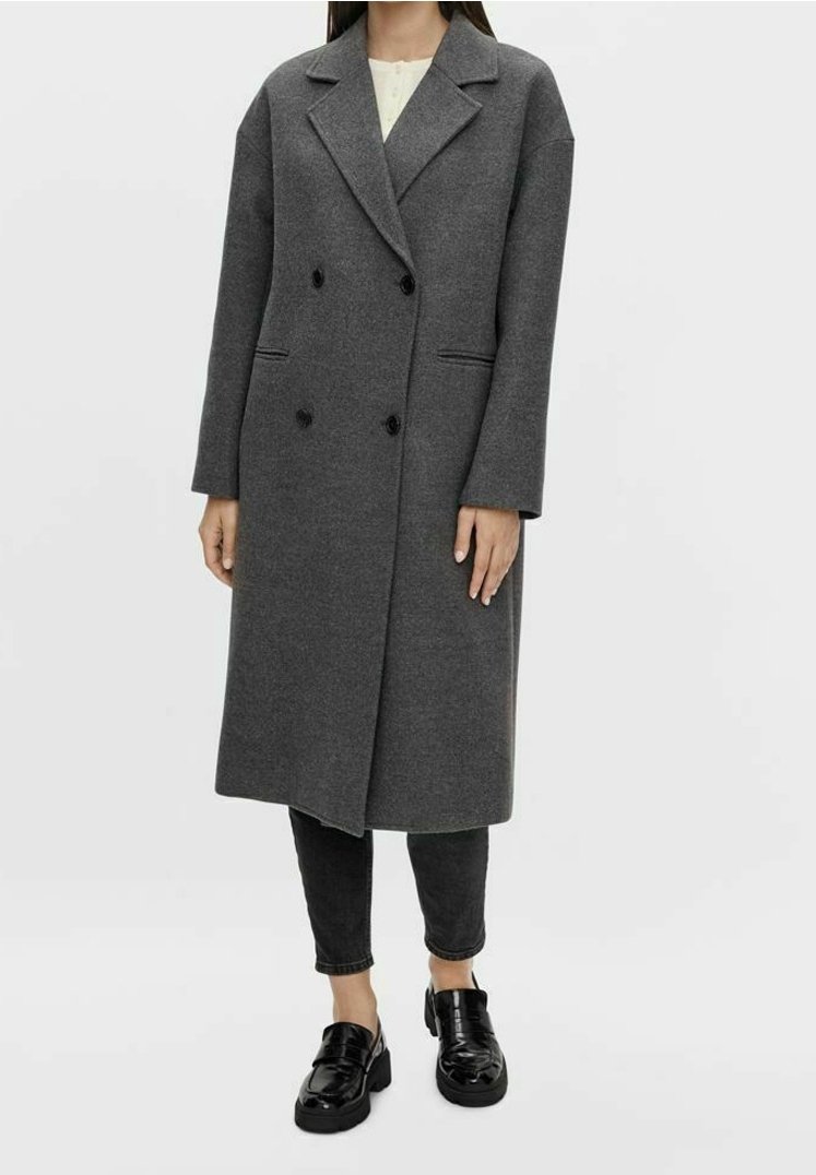 Personne portant un long manteau gris foncé à double boutonnage avec des boutons noirs, un pantalon noir et des mocassins noirs brillants sur un fond clair uni.