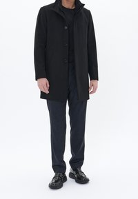 Mustaksi villakangastakiksi, jossa on korkea kaulus, nappilista ja kaksi sivutaskua, puettuna mustan paidan ja tummansinisten teksturoiden housujen päälle, yhdistettynä mustiin kenkiin.