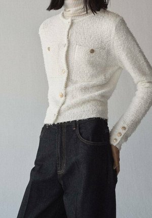 Hvid textured cardigan med knapper foran og guldknapper, båret over en hvid rullekrave, kombineret med høj taljede mørke jeans.