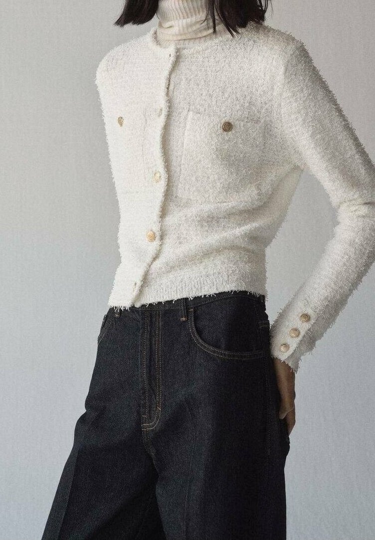Cardigan blanc texturé boutonné avec poches avant et boutons dorés porté sur un col roulé blanc, associé à un jean foncé taille haute.