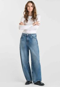 Femme debout, les bras croisés, portant un haut à manches longues blanc, un jean bleu ample et des chaussures noires, sur un fond uni.