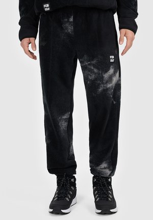 Osoba nosząca czarne i szare spodnie dresowe tie-dye z małym białym logo na lewej udzie oraz czarne buty trekkingowe z białymi podeszwami.