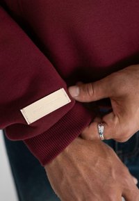 Una mano che sistema il polsino di un maglione bordeaux con un'etichetta beige che reca la scritta "PROIBITO," mostrando un anello d'argento sul pollice.