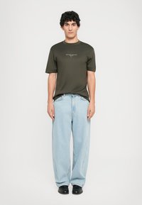 Olíva zöld, rövid ujjú, kerek nyakú póló, világos kék, oversized farmernadrággal párosítva. A modell fekete cipőt visel.