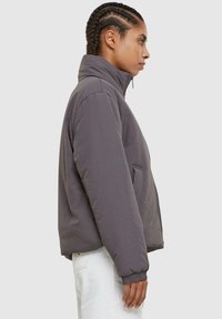 Grå, isolert jakke med høy krage, sidelommer, elastiske mansjetter og en glatt tekstur, vist på en modell i profil.